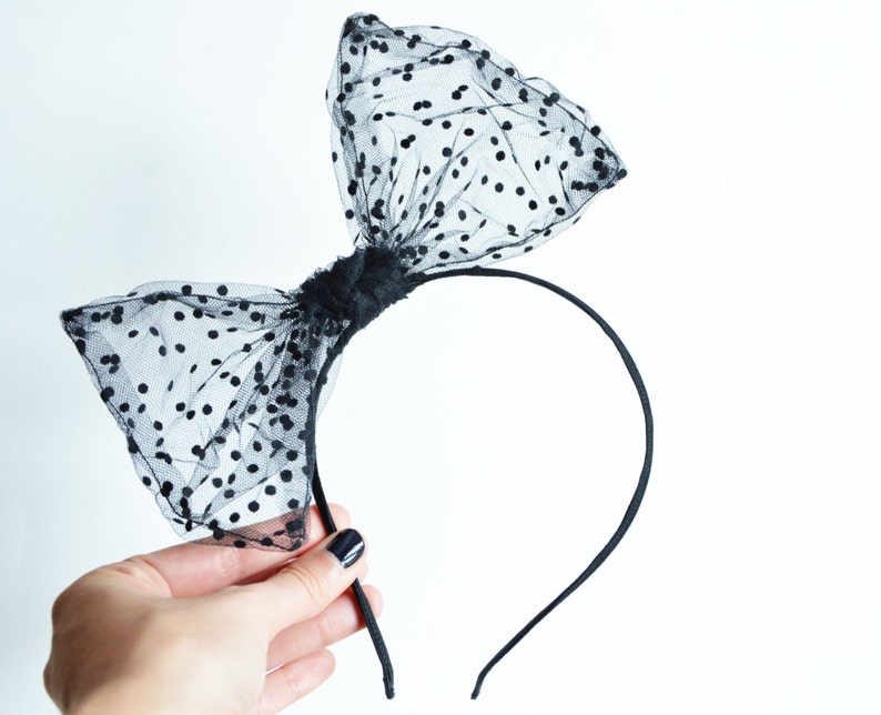 May include: Black headband featuring a large bow made of sheer black fabric with small, evenly spaced black polka dots. The bow is centered on a thin, black plastic headband, creating a stylish hair accessory.