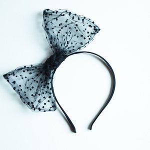 May include: A black headband with a large bow made of sheer fabric with black polka dots. The bow is centered on the headband, creating a stylish hair accessory. The headband is a simple, thin band.