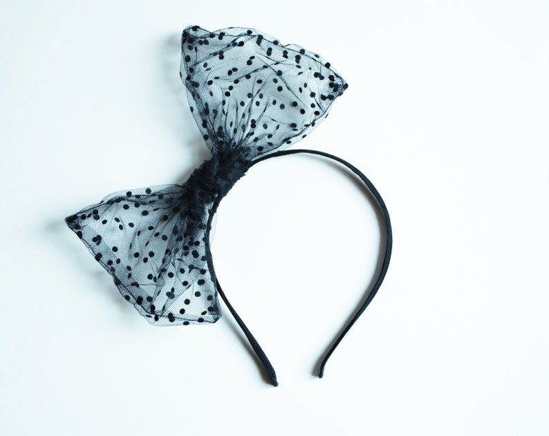 May include: A black headband with a large bow made of sheer fabric with black polka dots. The bow is centered on the headband, creating a stylish hair accessory. The headband is a simple, thin band.