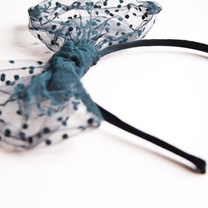 May include: A teal headband with a bow made of sheer fabric dotted with black spots. The bow is centered on a thin, black headband. The accessory is set against a white background.