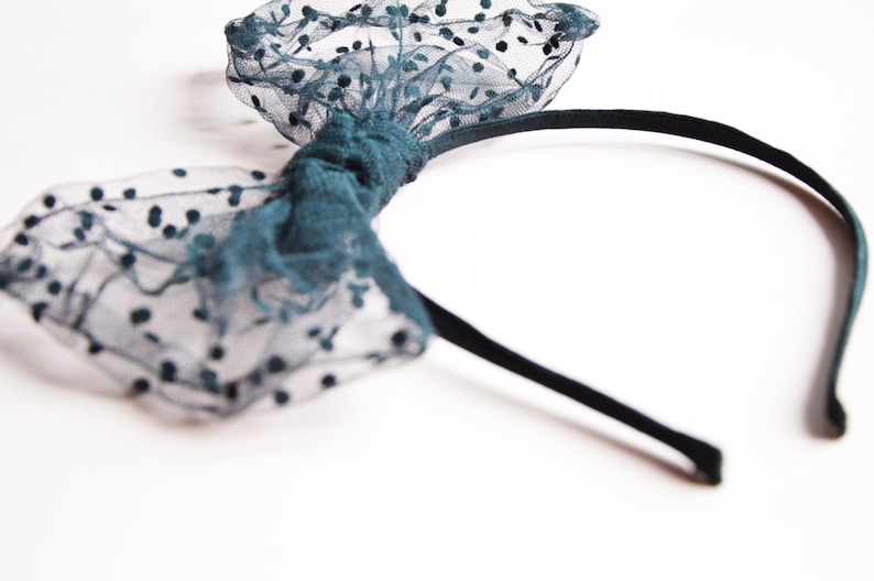May include: A teal headband with a bow made of sheer fabric dotted with black spots. The bow is centered on a thin, black headband. The accessory is set against a white background.