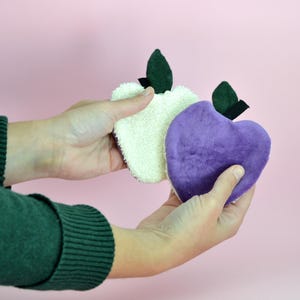 May include: Two fabric apple-shaped makeup remover pads. One pad is white and the other is purple.