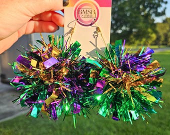 以下が含まれることがあります： マルディグラをテーマにしたイヤリングのペア。イヤリングは、緑、紫、金色のキラキラしたものでできており、球形に配置されています。イヤリングは金色のフックから吊り下げられています。背景には草地と木が含まれています。