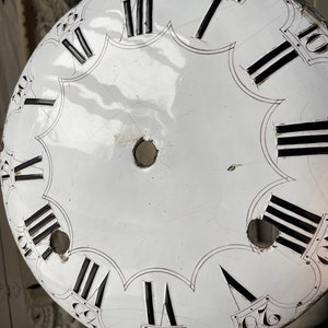 May include: A white clock face with black Roman numerals and minute markers. The face has a scalloped edge and two holes near the center. The clock face has visible cracks and signs of age.
