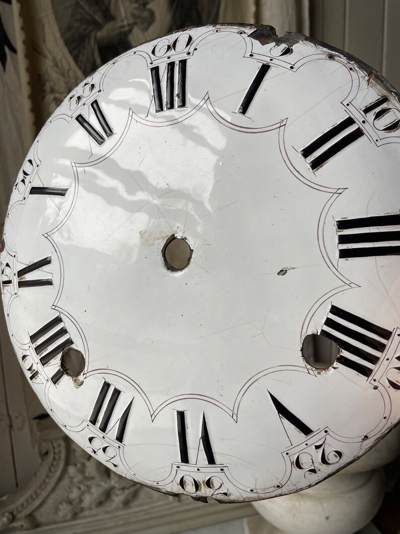 May include: A white clock face with black Roman numerals and minute markers. The face has a scalloped edge and two holes near the center. The clock face has visible cracks and signs of age.
