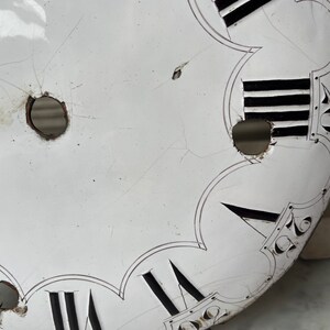 May include: Close-up of a vintage clock face with Roman numerals and black markings on a white background. The clock face has visible cracks and holes, suggesting age and wear. The design includes decorative elements.