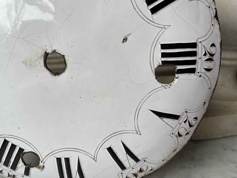 May include: Close-up of a vintage clock face with Roman numerals and black markings on a white background. The clock face has visible cracks and holes, suggesting age and wear. The design includes decorative elements.