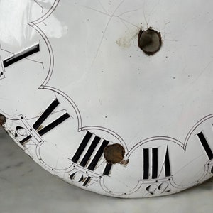 May include: Close-up of a vintage clock face with a white enamel finish. Black Roman numerals mark the hours, with minute markers around the edge. The face has visible cracks and two holes, suggesting age and use. A classic timepiece.