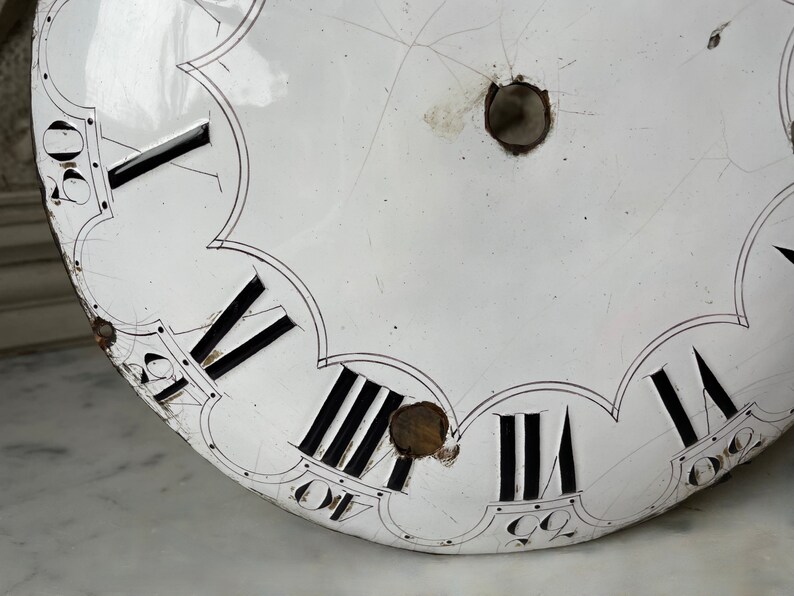 May include: Close-up of a vintage clock face with a white enamel finish. Black Roman numerals mark the hours, with minute markers around the edge. The face has visible cracks and two holes, suggesting age and use. A classic timepiece.