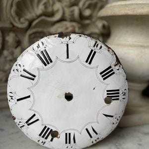 May include: Antique white clock face with black Roman numerals and minute markers. The clock face has a scalloped edge and a central hole. The face shows signs of age and wear, with some chipping and discoloration.