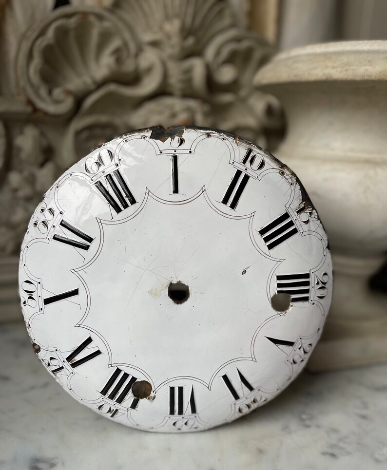May include: Antique white clock face with black Roman numerals and minute markers. The clock face has a scalloped edge and a central hole. The face shows signs of age and wear, with some chipping and discoloration.