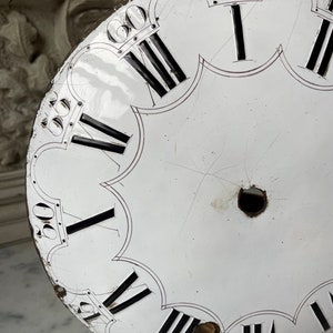 May include: A distressed white clock face with black Roman numerals and minute markers. The clock face has a scalloped edge and shows signs of age, including scratches and holes. The clock face is a vintage item.