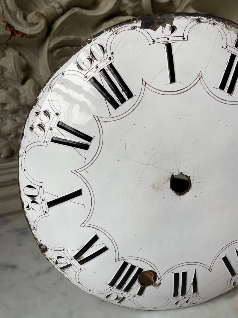 May include: A distressed white clock face with black Roman numerals and minute markers. The clock face has a scalloped edge and shows signs of age, including scratches and holes. The clock face is a vintage item.