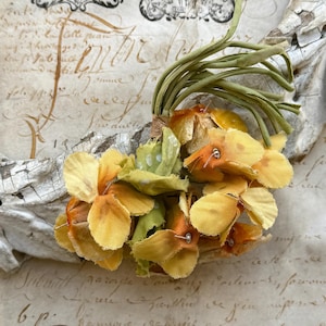May include: A cluster of yellow and orange fabric flowers with green stems and leaves. The flowers are attached with pins. The background features aged paper with handwritten script and decorative stamps.