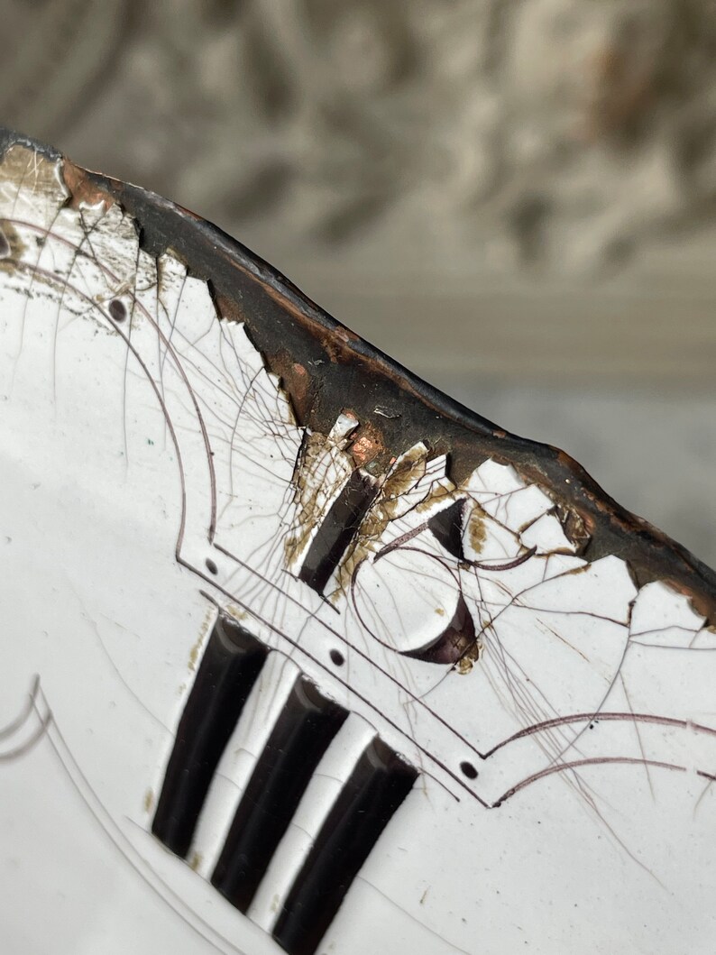 May include: Close-up of a white ceramic plate with black and brown accents. The plate features a cracked surface, black vertical stripes, and a decorative design with the number 16. The rim is dark brown.