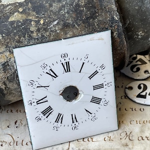 May include: A square, white clock face with black Roman numerals and minute markers. The face has a central hole and is cracked. The background includes a dark, textured object and a piece of paper with handwritten text.