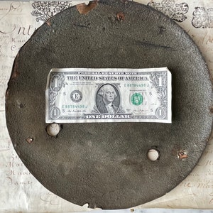 May include: A weathered, circular metal object with two holes, possibly a lid or plate. A one-dollar bill rests on top, providing a size reference. The object has a dark, textured surface and signs of wear. The background includes aged paper with handwritten script.