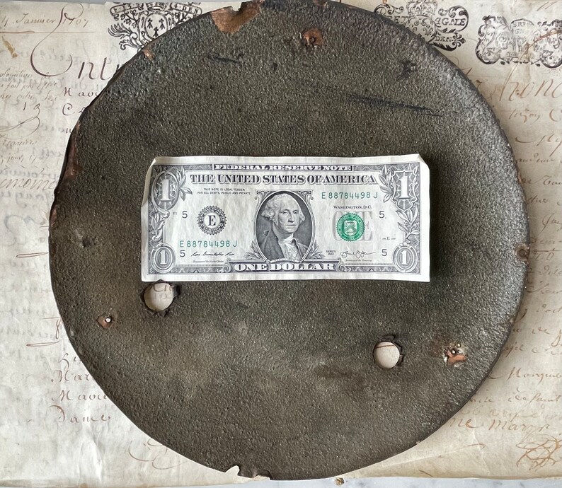 May include: A weathered, circular metal object with two holes, possibly a lid or plate. A one-dollar bill rests on top, providing a size reference. The object has a dark, textured surface and signs of wear. The background includes aged paper with handwritten script.
