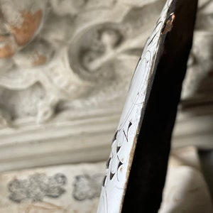 May include: Close-up of a decorative, elongated object with a white surface and intricate black floral patterns. The edge is dark brown with visible wear. The background features ornate, off-white architectural details.