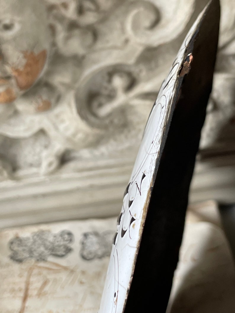 May include: Close-up of a decorative, elongated object with a white surface and intricate black floral patterns. The edge is dark brown with visible wear. The background features ornate, off-white architectural details.