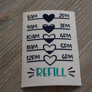 May include: A white sticker with black text and blue hearts. The sticker has a time schedule from 8 AM to 6 PM with a refill reminder at the bottom.