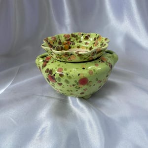 May include: A light green ceramic oil burner with a scalloped top dish and a rounded base featuring a speckled pattern of dark brown, orange, and red spots. The vessel has two small side handles and sits on three short, sturdy feet.