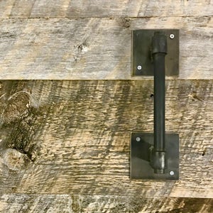 May include: A black metal door handle with a square base and a round, vertical bar. The handle is mounted on a rustic wooden wall.