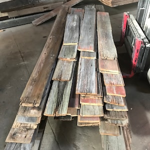 Puede incluir: Una pila de tablones de madera recuperada en varios tonos de gris, marrón y rojo. Los tablones de madera tienen un aspecto rústico y desgastado y son perfectos para proyectos de artesanía.