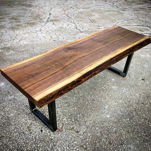 May include: A long, rectangular wooden bench with a live edge top and black metal legs. The wood is a rich brown color with a natural, rustic finish.