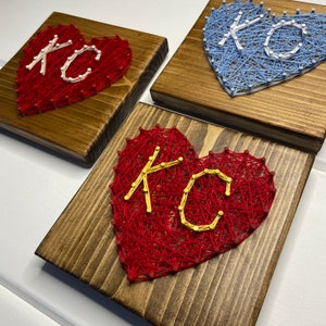May include: Three wooden plaques with string art hearts. The hearts are red and blue and have the letters "KC" in white and yellow thread.