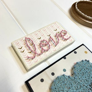 May include: A pink string art piece with the word "Love" spelled out on a white background with a pink and gold chevron pattern.