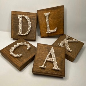 May include: Five wooden squares with the letters D, L, C, A, and K spelled out in white string art. The letters are strung on nails hammered into the wood.