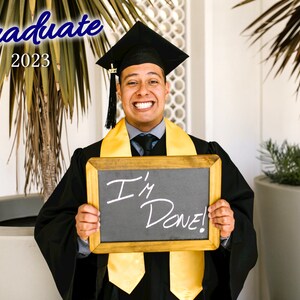 May include: A person wearing a black graduation cap and gown, holding a chalkboard with the words "I'm Done!" written on it. The person is smiling and has a yellow graduation sash.