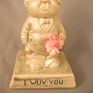 May include: A ceramic figurine of a person with a large head and a goofy expression. The figurine is holding a pink flower and is standing on a base that says "I WUV YOU".