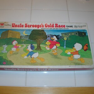 May include: A vintage board game box featuring Uncle Scrooge and Donald Duck characters from Disney's comics. The box is green, yellow, and brown with a cartoon illustration of the characters. The game is called "Uncle Scrooge's Gold Race" and is for 2 to 4 players, ages 7 to 10.