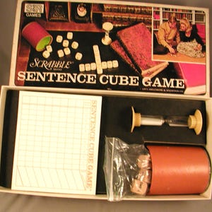May include: A Scrabble Sentence Cube Game box set with a brown and white game board, a red and green dice cup, white dice, and a white hourglass. The box lid features a black background with the words "Scrabble Sentence Cube Game" in white.