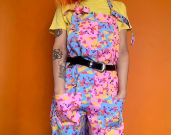 May include: A person wearing a pair of blue, pink, and yellow patterned overalls with a black belt. The overalls have large pockets on the front. The person is wearing a yellow t-shirt and has orange hair.