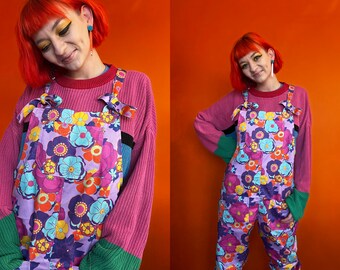 May include: A person wearing a pink sweater with a floral print overall jumpsuit. The overall jumpsuit has a purple background with pink, blue, yellow, and orange flowers.