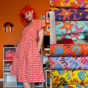 May include: A red and white checked dress with a heart pattern. The dress has short sleeves and a gathered waist. Beside the dress are stacks of colourful fabric with starburst and abstract designs, along with a spool of white thread.