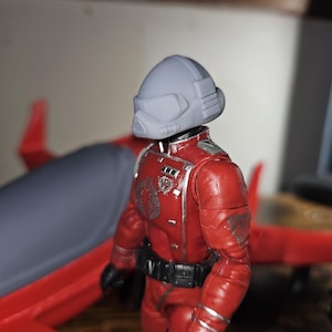 Puede incluir: Una figura de acción roja y plateada con un casco gris con un logotipo de Cobra en el pecho. La figura está de pie junto a un avión rojo y gris.