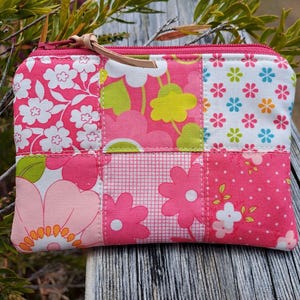 May include: A patchwork zipped pouch with a variety of pink floral patterns. The pouch has a leather pull tab and is made of quilted fabric. The design includes white, green, and yellow flowers.
