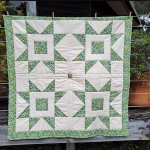 May include: A handmade quilt with a cream-colored background and green floral and geometric patterns. The quilt features squares, triangles, and a green floral border. It is hanging on a clothesline outdoors.