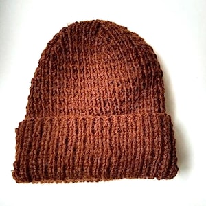May include: A brown knit beanie hat with a folded brim. The hat is made of a textured, ribbed knit material. The hat is a solid color and is displayed on a white background.