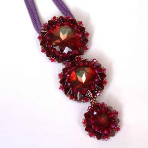 May include: A necklace featuring three red crystal beads, each surrounded by smaller red beads. The necklace is made with a purple cord.