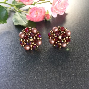 Puede incluir: Un par de pendientes redondos de cuentas con un esquema de color oro rosa y burdeos. Los pendientes están hechos con pequeñas cuentas redondas y cristales.