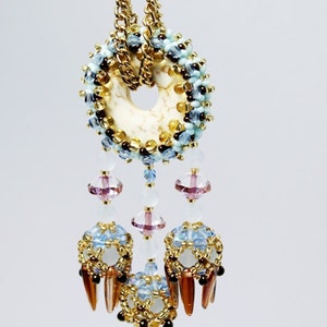 May include: A gold chain necklace with a white and blue beaded pendant. The pendant has a round, open centre and is decorated with small, clear crystals. The pendant is attached to a chain with three strands of beads, each ending in a small, clear crystal.