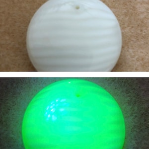 Könnte beinhalten: Eine weiße runde Perle mit einem Loch oben. Die Perle leuchtet unter Schwarzlicht grün.