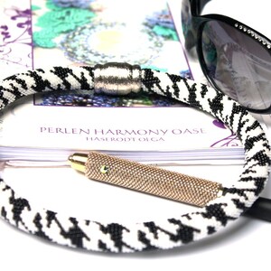 May include: A black and white beaded necklace with a silver toggle clasp. The necklace is lying on a book with the text "Perlen Harmony Oase Hase root Olga".