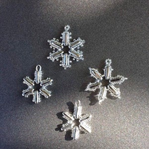 Könnte beinhalten: Vier silberne Schneeflocken-Ornamente in verschiedenen Größen. Die Schneeflocken sind aus Draht gefertigt und haben ein filigranes, kompliziertes Design.