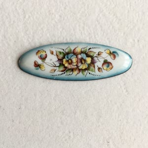 May include: An oval-shaped enamel pin with a blue background and a hand-painted floral design in shades of yellow, pink, and green.